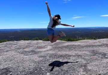 A girl is jumping high on top of Bald Rock, which is a giant granite monolith. There is countryside and blue sky in the background.