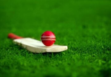 A red cricket ball balancing on a cricket bat lying on the green grass.