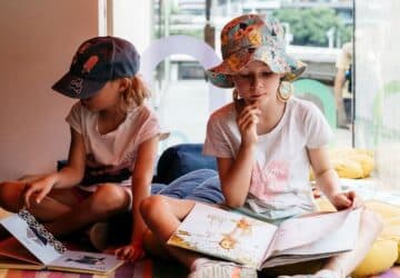 Summer reading club image of two girls reading books in the sun with hats on.